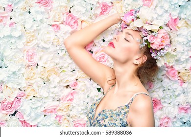 Beautiful Romantic Young Woman In A Wreath Of Flowers Posing On A Background Of Roses. Inspiration Of Spring And Summer. Perfume, Cosmetics Concept.