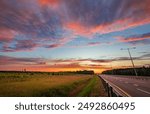 A beautiful rural highway at sunset with a colorful sky, clouds, grassy field, and trees creating a peaceful scene