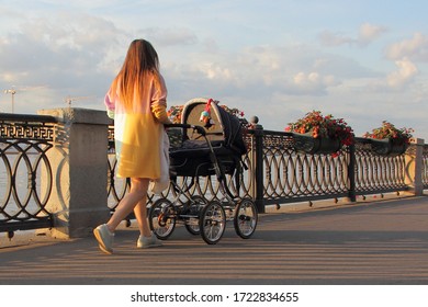 Beautiful Russian Young Mother Woman In A Light Dress Walks With A Baby Stroller On Moscow River Embankment Park Alleyway On A Sunny Summer Day With Sun Shine, Maternal Care