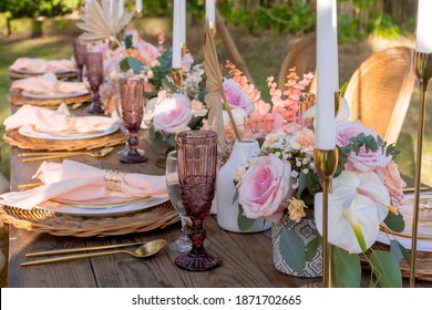 A beautiful rustic table setup at an outdoor garden wedding. White tall candles and pastel rose and tulip tabletop bouquet. Late afternoon shot. Open-air banquet, outside