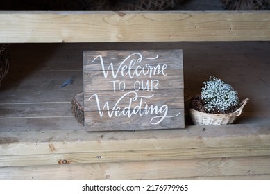 Beautiful Rustic Welcome To Our Wedding Sign. White Writing On A Dark Wooden Board, With Flowers Beside.