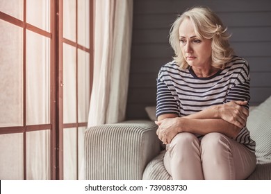 Beautiful sad senior woman is looking downward while sitting on couch at home