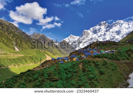 Beautiful scenic view of green meadows, snowy mountains with blue sky, shops, hotels and crowd of Shiva devotees on the way to Kedarnath Temple Trek