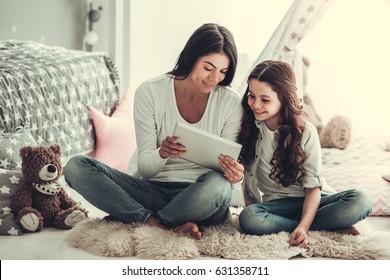 Beautiful school girl and her mom are using a digital tablet and smiling while sitting in girl's room at home