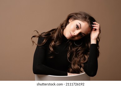 Beautiful seductive young european woman portrait. Girl wear black sweater with beautiful face and hair sitting on chair looking at camera. Isolated on brown background
