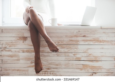 Beautiful seductress. Cropped image of gorgeous young African woman with beautiful legs sitting on the window sill with laptop laying near her 