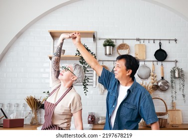 Beautiful Senior Asian Couple Is Dancing And Smiling While Cooking Together Kitchen. Full Length Energetic Middle Aged Family Couple Dancing To Disco Music In Kitchen. Happy Old Mature People Happy.