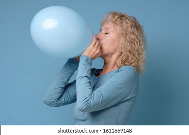 Beautiful senior blonde woman preparing for party blowing up blue balloon