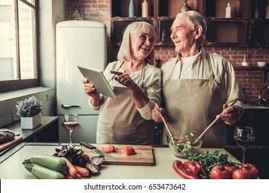 Beautiful senior couple in aprons is using a digital tablet, talking and smiling while cooking together in kitchen - Powered by Shutterstock