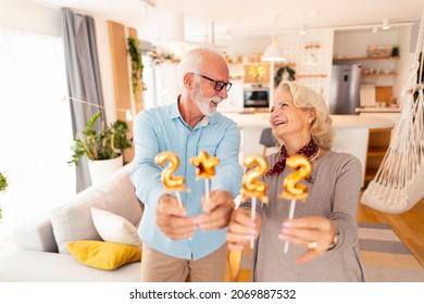 Beautiful Senior Couple Holding Golden Balloons Shaped As Numbers 2022, Representing The Upcoming New Year While Spending Winter Holiday Season Together At Home