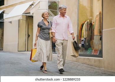 Beautiful Senior Couple Holding Hands Walking In Luxury Shopping Street Carrying Bags, Vacation Trip, Outdoors. Healthy Mature Consumer People On City Break Travel, Leisure Recreation Lifestyle.