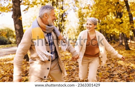 Beautiful senior couple laughing and running together in park on autumn day, two pensioners wife and husband enjoying happy life moments on retirement on walk outdoors