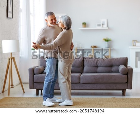 Beautiful senior couple in love looking at each other with tenderness while dancing together in living room at home, two pensioners wife and husband enjoying happy life moments on retirement