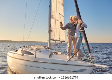 Beautiful senior couple making selfie on smartphone while standing on the side of sailboat or yacht deck floating in sea, they are hugging and smiling at camera
