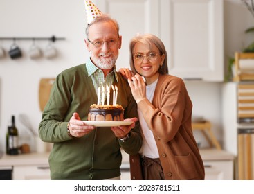 Beautiful Senior Couple In Party Hats Celebrating Birthday And Anniversary Together At Home, Holding Chocolate Cake, Hugging And Smiling While Standing In Modern Kitchen. Family Celebration Concept