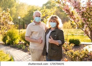 Beautiful Senior Couple Showing Thumbs Up And Wearing Medical Mask To Protect From Coronavirus Outside In Spring Or Summer Nature