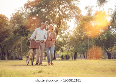 Beautiful Senior Couple Walking Their Bike Along Happily Talking In Park.mature Couple In Summer Park.Elderly Man And Old Woman With Bicycles Outside In Spring Nature.