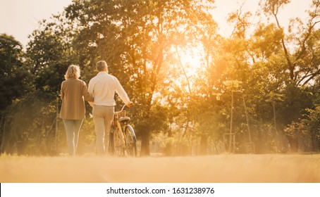 Beautiful Senior couple walking their bike along happily talking in park.Elderly man and old woman with bicycles outside in spring nature.Happy senior couple  holding hands while walking at sunset.