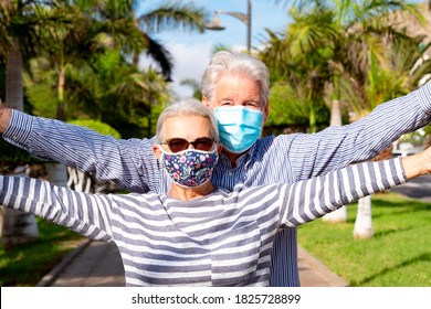 Beautiful senior couple with white hair having fun in public park with a sunset light, with raised arms, wearing surgical mask due to coronavirus