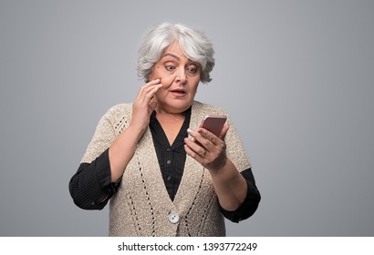 Beautiful senior female looking at modern smartphone with astonished face expression while standing on gray background - Powered by Shutterstock