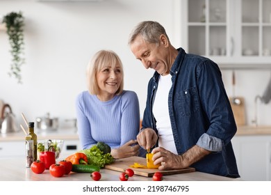 Beautiful Senior Spouses Having Conversation And Smiling While Cooking At Home, Cheerful Pretty Elderly Woman Supporting Her Husband Preparing Healthy Dinner, Cutting Vegetables, Copy Space