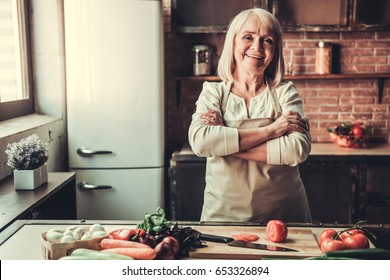 Beautiful Senior Woman In Apron Is Looking At Camera And Smiling While Standing With Crossed Arms In Kitchen