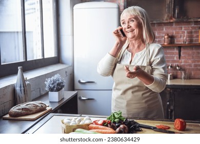 Beautiful senior woman in apron is talking on the mobile phone and smiling while cooking in kitchen - Powered by Shutterstock