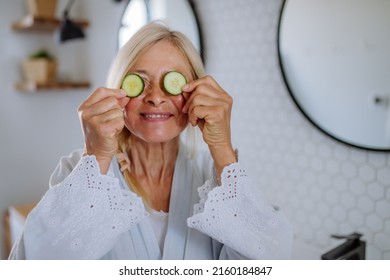 Beautiful Senior Woman In Bathrobe Applying Cucumber Face Mask In Bathroom, Skin Care Concept.