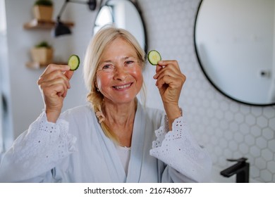 Beautiful Senior Woman In Bathrobe Applying Cucumber Face Mask In Bathroom, Skin Care Concept.