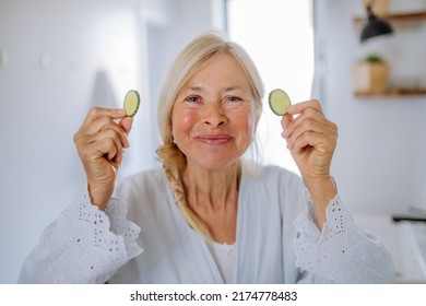 Beautiful Senior Woman In Bathrobe Applying Cucumber Face Mask In Bathroom, Skin Care Concept.