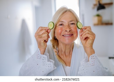 Beautiful Senior Woman In Bathrobe Applying Cucumber Face Mask In Bathroom, Skin Care Concept.