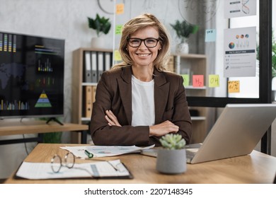 Beautiful Senior Woman With Blond Hair Looking At Camera While Sitting At Desk With Papers And Modern Laptop. Business Lady In Stylish Formal Suit Posing At Own Office