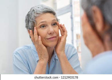 Beautiful senior woman checking her face skin and looking for blemishes. Portrait of mature woman massaging her face while checking wrinkled eyes in the mirror. Aging process concept.