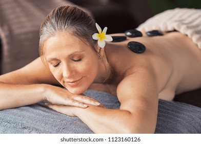 Beautiful Senior Woman With Eyes Closed Hot Stone Massage At Salon Spa. Portrait Of Mature Woman Relaxing During Lastone Therapy. Senior Lady Lying On Spa Table With Black Lava Stones On Back.