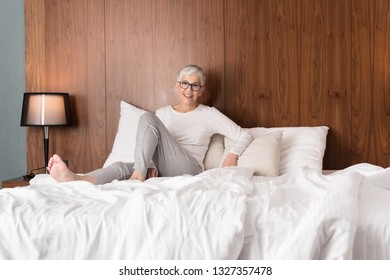 Beautiful Senior Woman With Glasses Sitting On A Comfy Bed And Getting Ready For Sleep In Her Bright Modern Bedroom, Night Routine And Bedtime Concept