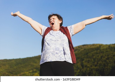 Beautiful senior woman holding both hands in the air while yelling with mouth wide open with the blue sky in the background - Cute old lady feeling energized outside - Elderly person feeling release - Powered by Shutterstock