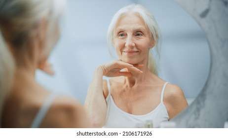 Beautiful Senior Woman Morning Bathroom Routine, Looks Into Mirror Touches Her Neck With Sensual Movement Of Hands. Elderly Female Natural Beauty. Dignity And Grace In Old Age. Concept For Skin Care