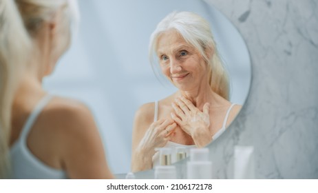 Beautiful Senior Woman Morning Bathroom Routine, Looks Into Mirror Touches Her Neck With Sensual Movement Of Hands. Elderly Female Natural Beauty. Dignity And Grace In Old Age. Concept For Skin Care