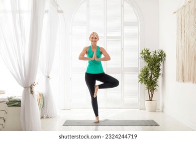 Beautiful Senior Woman Practicing Yoga At Home, Standing In Tree Pose, Happy Elderly Lady In Activewear Exercising Indoors In Light Room, Smiling Female Enjoying Healthy Lifestyle, Copy Space