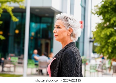 Beautiful Senior Woman With Short Gray Hair Outdoors Against City Background. Waist Up Portrait Of Confident Attractive Older Female In The City