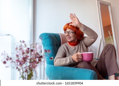 Beautiful Senior Woman Sitting In An Armchair By The Window, Drinking Tea And Waving To Neighbors Through The Window