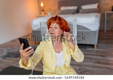 Beautiful senior woman sitting on a yoga mat, putting on headset, relaxing after doing yoga, listening to the music