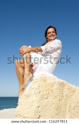 Beautiful senior woman sitting relaxed, joyful and happy on rock at beach, enjoying holiday, retirement at sea, isolated with ocean and blue sky as background and copy space.