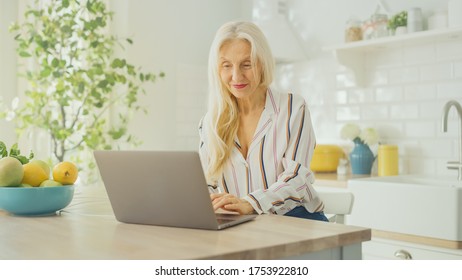 Beautiful Senior Woman Using Laptop Computer In A Sunny Kitchen. She Smiles. Grandmother Is Connected With Children Via Internet And Social Media. Pensioner Working From Home.