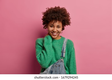 Beautiful sensitive woman with curly hair looks at interesting thing, touces neck and smiles happily, hears nice idea, wears green sweater, isolated on pink background. Human face expressions