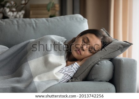 Beautiful serene woman sleeping on comfy sofa under warm cozy plaid in modern living room. Healthy daytime nap, tiredness relief, repose and relaxation during day, lazy weekend at home. Rest concept