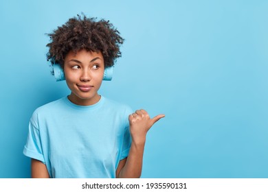 Beautiful serious Afro American woman with dark skin dressed in casual t shirt points thumb away gives recommendation listens music via headphones shows copy space against blue studio background - Shutterstock ID 1935590131