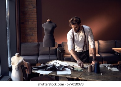 Beautiful serious tailor with glasses in white shirt with brown leather suspenders working near wooden table with threads, apron and scissors in amazing atelier with antique furniture and mannequin