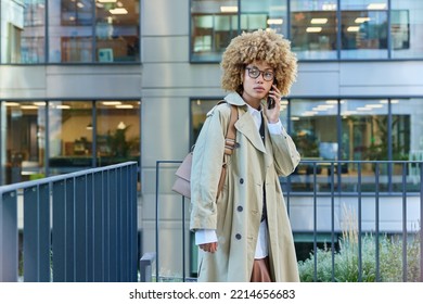 Beautiful Serious Young Woman With Curly Bushy Hair Has Telephone Conversation Focused Somewhere Enjoys Leisure Time Wears Transparent Spectacles And Beige Coat Poses Against Urban Building.