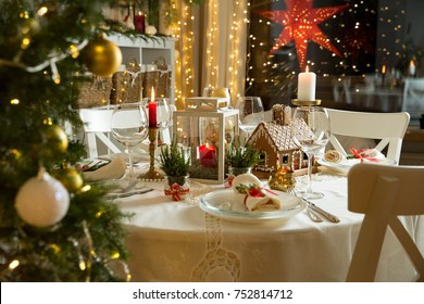 Beautiful served table with decorations, candles and lanterns. Little gingerbread house with glaze on white tablecloth. Living room decorated with lights and Christmas tree. Holiday setting